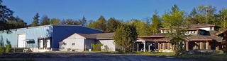 A blue warehouse next to a multi-story wooden building under a clear blue sky with green trees.