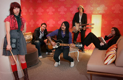 A five-member band posing in a room with red clover-patterned wallpaper, holding various musical instruments.