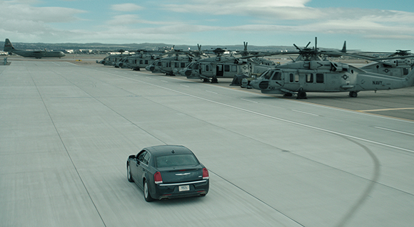 A dark sedan drives on a concrete airfield past a row of parked military helicopters and a large plane.