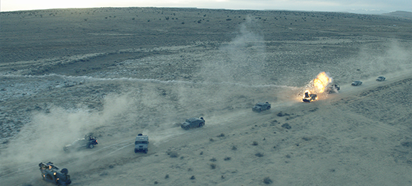 Military vehicles drive through a dusty desert landscape as one vehicle in the convoy explodes.