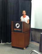 A woman in a white shirt and orange necklace stands behind a dark podium with a circular logo.