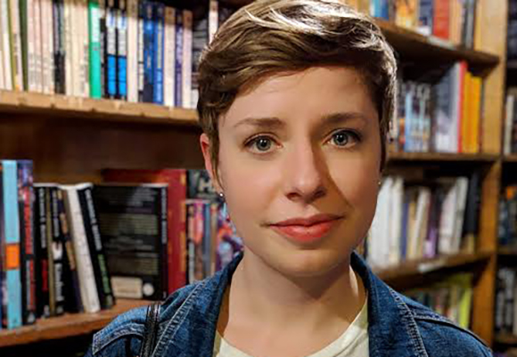 A person with short brown hair and blue eyes looking forward in a library setting.