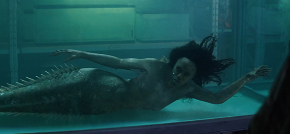 A mermaid with dark hair and a long scaly tail swims inside a large, dimly lit water tank.