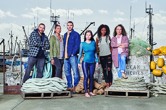 Six diverse people pose together on a dock with fishing boats and nets in the background.