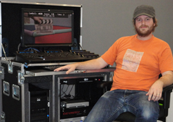 A man in an orange shirt and cap sits beside a large black equipment rack with a monitor and control panel.