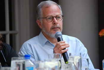A man with graying hair and glasses holds a microphone while speaking at a table.