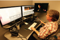 A person in a plaid shirt sits at a desk using a keyboard and mouse with three large computer monitors.
