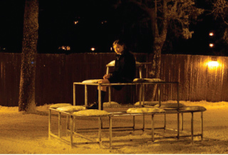 A person sits on a low, circular metal frame structure outdoors at night near a wooden fence.