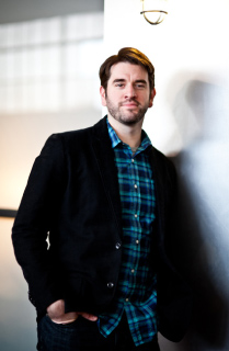 A man with short brown hair wearing a black blazer and blue plaid shirt leaning against a light-colored wall.