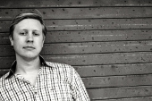 A man in a checkered shirt stands in front of a dark, horizontal wooden plank wall.