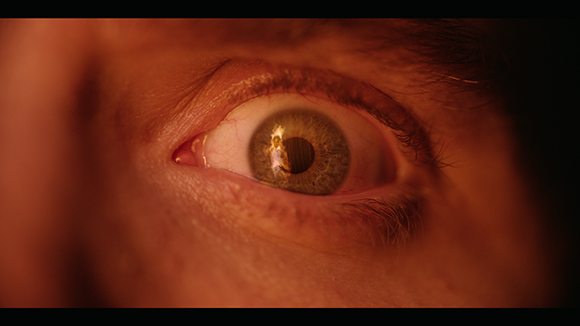 Extreme close-up of a wide-open human eye under warm, reddish lighting.