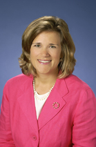 A smiling woman with blonde hair wearing a pink blazer and a pearl necklace against a blue background.