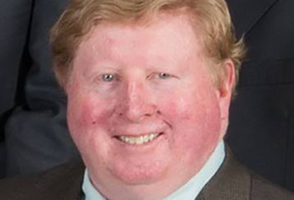 A smiling man with light reddish hair and fair skin wearing a light blue shirt and brown jacket.