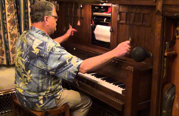 A man in a patterned shirt sits at a wooden player piano, reaching for the controls.