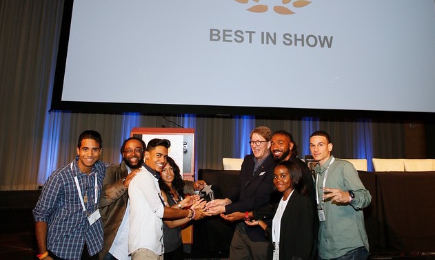 A diverse group of people smiling and posing together on a stage in front of a screen reading BEST IN SHOW.