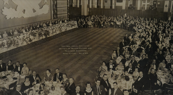 A large crowd of people seated at banquet tables around a large, empty wooden dance floor in a grand hall.