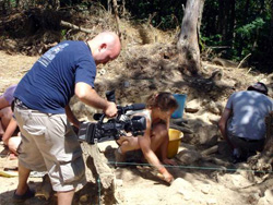 A man in a blue shirt operates a professional video camera toward a woman sitting in a dirt excavation site.