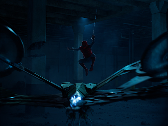 A figure in a red suit swings on a web above a large mechanical structure in a dark, blue-lit warehouse.