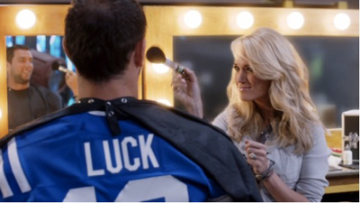 A blonde woman uses a makeup brush on a man wearing a blue jersey with 'LUCK' on the back.
