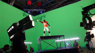 A person in an orange jersey stands on a raised platform in front of a large green screen studio backdrop.