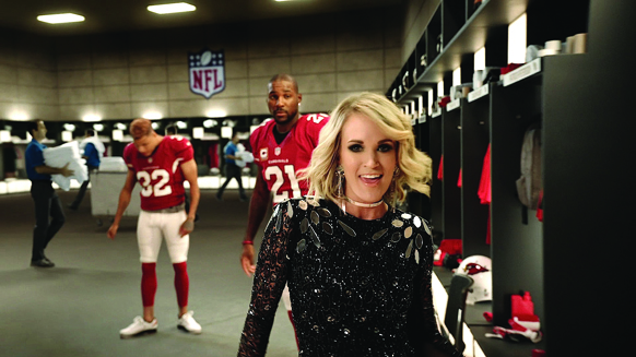 A woman in a black sequined dress smiles in an NFL locker room with players in red jerseys behind her.