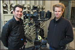 Two men pose next to a large professional cinema camera mounted on a tripod outdoors.