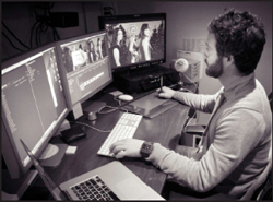A man sits at a desk using a keyboard, mouse, and stylus with three computer monitors.