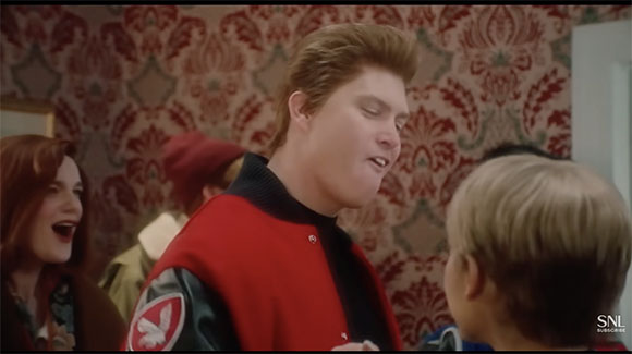 A young man with voluminous hair in a red jacket looks toward a child in front of patterned wallpaper.