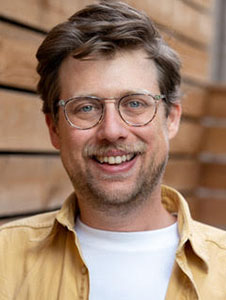 A smiling man with wavy brown hair, glasses, and a mustache wearing a yellow shirt.