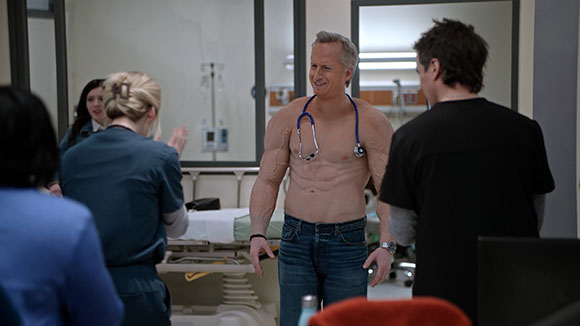 A shirtless man in blue jeans smiles while wearing a stethoscope around his neck in a hospital room.