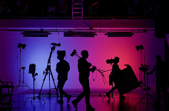 Silhouettes of a film crew and camera equipment against a blue and red lit studio background.