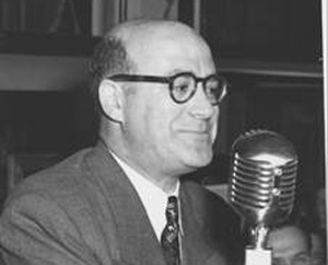 A man in a suit and glasses speaks into a vintage silver microphone.