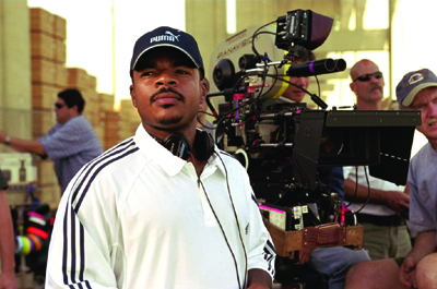 A man in a white polo and blue cap stands before a large professional film camera on a set.