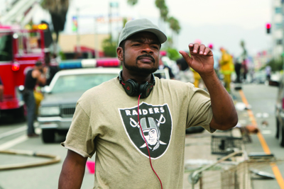 A man wearing a grey cap and a Raiders t-shirt with headphones around his neck stands on a street.