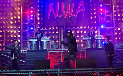 Performers on a stage with bright pink and yellow lights and a large N.W.A sign in the background.