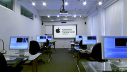 A classroom filled with rows of desks, computer monitors, and black chairs under bright ceiling lights.
