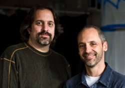 Two men with facial hair smiling in a dimly lit indoor setting.