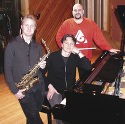 Three men pose with a saxophone and a grand piano in a room with wood-paneled walls.