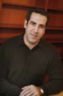 A man with dark hair wearing a black polo shirt sits before wooden bookshelves.