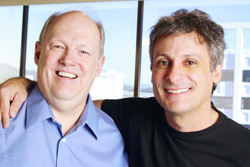 Two smiling men posing together indoors in front of a large window.