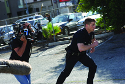 A police officer in dark uniform runs past a person filming with a professional video camera.