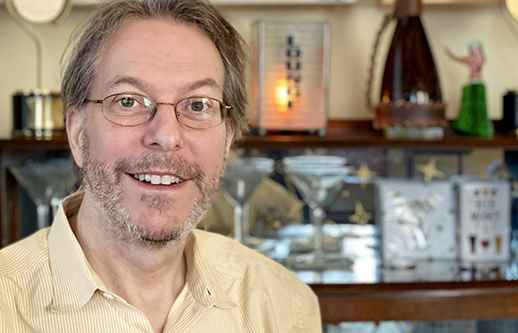 A man with glasses and a salt-and-pepper beard smiles in a bar setting.