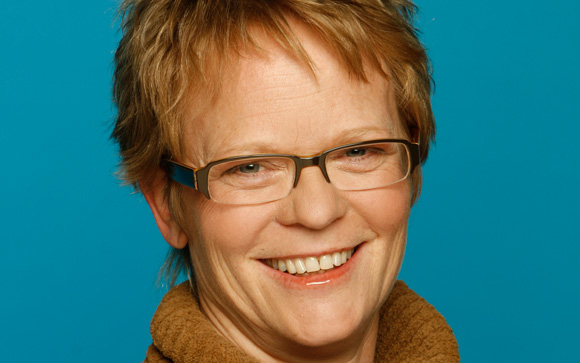 Smiling woman with short reddish-blonde hair and rectangular glasses against a blue background.