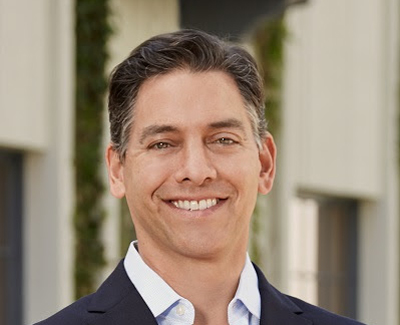 A smiling man with dark hair wearing a light blue shirt and dark blazer.