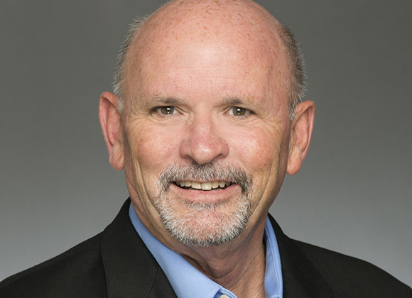 A smiling middle-aged man with light skin, a bald head, and a graying goatee wearing a black blazer and blue shirt.
