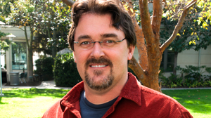 A man with dark hair, glasses, and a goatee wearing a red shirt, smiling in front of trees.
