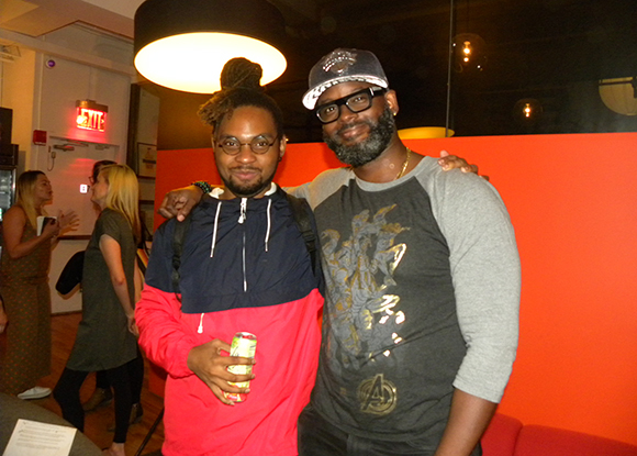 Two men smiling and posing for a photo in a dimly lit room with a red wall.