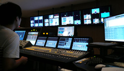 A person sits before a large video mixing console and multiple monitor screens in a dark control room.