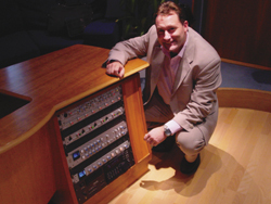 A man in a tan suit kneels beside a wooden audio equipment rack in a studio.