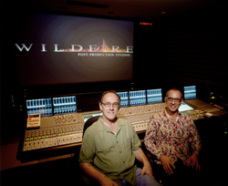 Two men sit behind a large audio mixing console in a dark studio with a screen in the background.